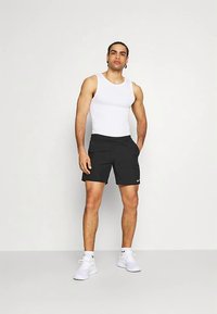 Schwarze Sportshorts mit Nike-Logo, kombiniert mit einem weißen Tanktop. Modell steht in weißen Sneakers und präsentiert eine taillierte Silhouette.