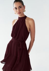 Bordo halter tipo suknelė, pagaminta iš plaukuojančios medžiagos, su daugiapakopiu raukšlėtu sijonu ir surišuotu liemeniu. Papuošta didelėmis auksinėmis apyrankėmis.