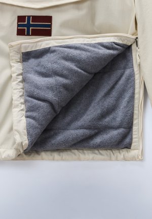 L'intérieur de la veste beige montre une doublure en polaire grise, avec un patch rouge foncé comportant un motif de drapeau bleu et blanc à l'extérieur.