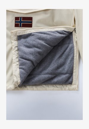 Interior de una chaqueta beige que muestra un forro de felpa gris, con un parche de color rojo oscuro que presenta un diseño de bandera azul y blanca en el exterior.