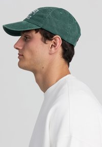 Cappellino da baseball in cotone verde scuro con visiera leggermente curvata e lettering ricamato. Indossato da una persona con una felpa bianca, vista di lato.