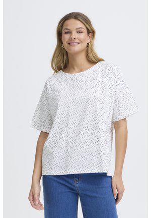 Femme souriante aux longs cheveux blonds portant un haut blanc à manches courtes avec de petits points noirs et un jean bleu, debout devant un fond uni.