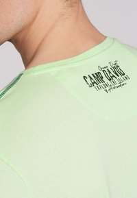 Camiseta de algodón verde claro con cuello redondo, detalles cosidos y un logo impreso cerca de la parte posterior del cuello.