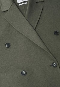 Manteau en laine vert foncé à double boutonnage avec de larges revers et quatre boutons noirs, montrant le col et les détails supérieurs à l'avant.