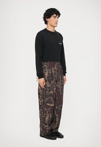 Zwarte long-sleeve shirt met het "Nohilite" logo, gecombineerd met losse bordeauxrode en bruine gebloemde broeken. Zwarte schoenen maken de outfit compleet.