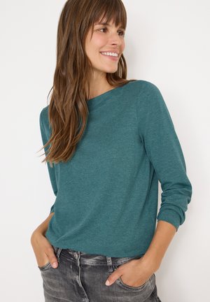 Maglietta a maniche lunghe di colore turchese realizzata in tessuto morbido e leggero, con un ampio scollo e polsini arrotolati, abbinata a jeans grigi strappati.