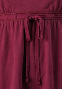 Bordeaux jurk met een gathered taille en een stoffen strik, gemaakt van zacht materiaal, met subtiele textuurdetails en contrasterende stiksels.