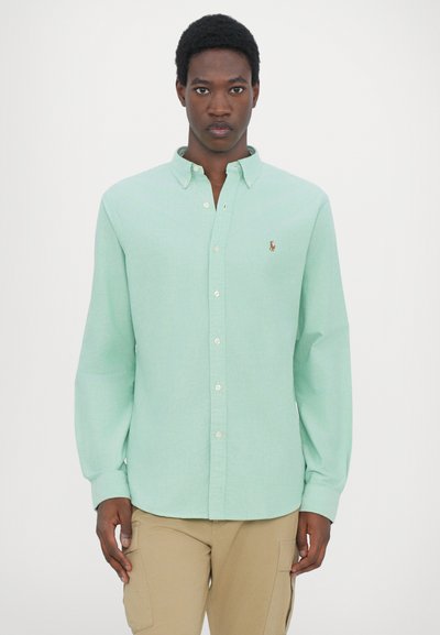 Chemise boutonnée vert clair avec col et manches longues, présentant un petit logo brodé sur la poitrine gauche ; texture de tissu lisse.