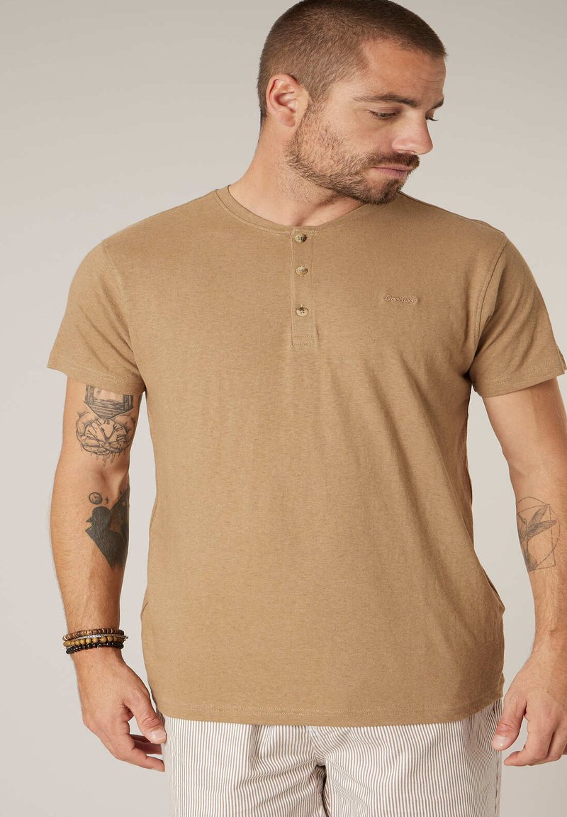 Homme aux cheveux courts et à la barbe, portant un t-shirt henley beige et un pantalon rayé, montrant des tatouages sur les deux bras et des bracelets au poignet gauche.