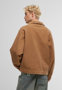 Braune Baumwolljacke mit lockerem Schnitt, großem Schalkragen und sichtbaren Nahtdetails. Getragen mit hellblauen Jeans.