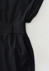 Zwarte kledingstuk van een gladde, lichte stof. Het heeft een gedefinieerde taille met een zakdetail en lichtgeplooide mouwen.