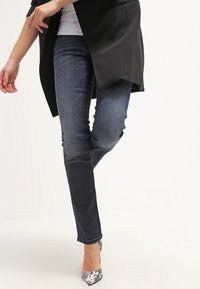 Jeans de mezclilla oscura con un corte slim, combinados con un blazer negro. El calzado presenta tacones con estampado de serpiente en plata y negro.