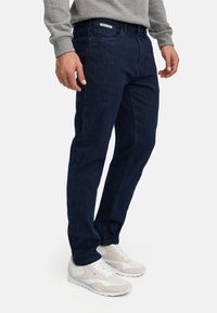 Homme portant un jean bleu foncé, une chemise grise à manches longues et des baskets blanches, debout devant un fond uni.