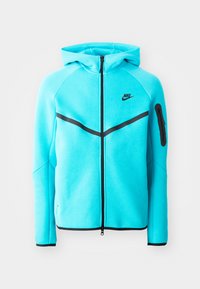 Tirkīza Nike kapuce ar pilnu rāvējslēdzēju, melniem akcentiem uz pleciem un kabatām, kā arī mazu logotipu uz krūtīm. Izgatavota no maigām, teksturētām audumam.