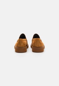 Zapatos de ante marrón tipo slip-on con punta redondeada, detalle de costura sutil y una suela de goma gruesa, vistos desde la parte trasera.