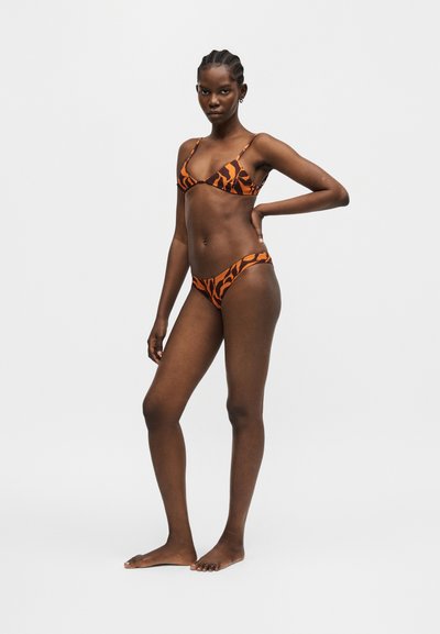 Γυναίκα όρθια ξυπόλητη με πορτοκαλί και καφέ μοτίβο μαγιό, με το ένα χέρι στη μέση και ουδέτερη έκφραση μπροστά σε απλό φόντο.