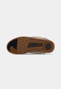 Sneaker sole featuring a textured rubber surface, predominantly beige with black accents, and an embossed logo. Designed for grip and durability.