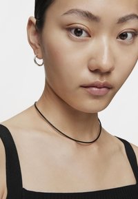 Collana choker nera con una finitura liscia, indossata con un top nero a coste. La modella indossa anche un piccolo orecchino a cerchio in argento. Visione ravvicinata.