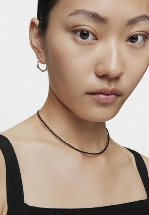 Collana choker nera con una finitura liscia, indossata con un top nero a coste. La modella indossa anche un piccolo orecchino a cerchio in argento. Visione ravvicinata.