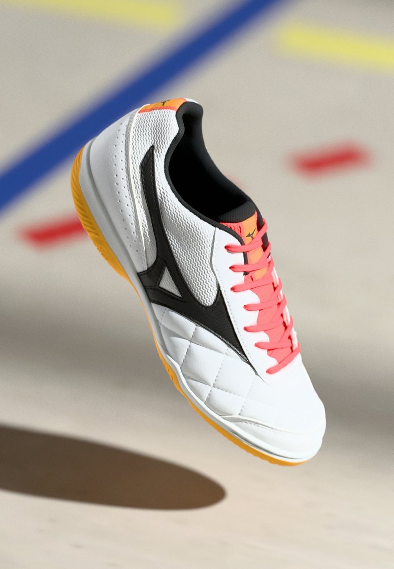 White athletic shoe with a quilted pattern, black accents, and bright pink laces. Yellow sole, synthetic material with textured detailing.