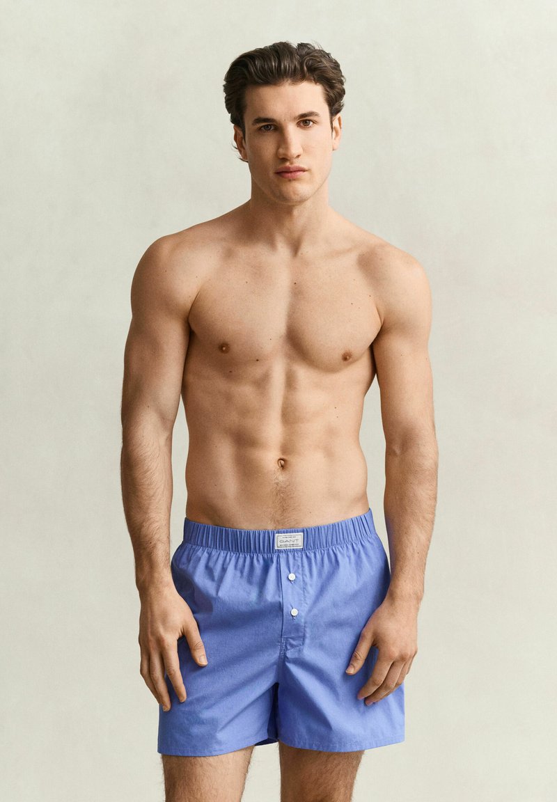 Jeune homme aux cheveux courts et foncés portant un boxer bleu, debout devant un fond clair uni.