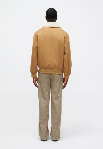 Καφέ φλις μπουφάν με γιακά από γούνα, ριμπ μανσέτες και ζωνάκι, σε συνδυασμό με ανοιχτό καφέ υφασμάτινο παντελόνι καμπάνα.
