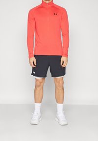 Haut de sport rouge à manches longues avec col zippé, associé à un short sombre et des baskets blanches. Présente un logo de marque noir sur le t-shirt.
