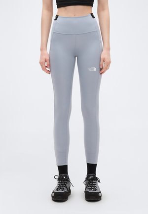 Γυναίκα που φοράει γκρι ψηλόμεσα κολάν The North Face, μαύρες κάλτσες και γκρι-μαύρα ορειβατικά παπούτσια, στέκεται μπροστά σε λευκό φόντο.