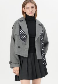 Giacca grigia a taglio corto in materiale simile al feltro, con una sciarpa a pois in accento. Il design include bottoni e maniche strutturate. Sotto, un top nero aderente e una gonna plissettata.