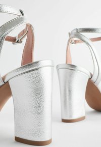 Hõbedased kõrged sandaalid metallilise viimistlusega, millel on mitu rihma ja massiivne konts. Siseosa on hele roosa ja sile tekstuur.