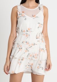 Weißes Blumenkleid mit Rüschenträgern, transparentem Ausschnitt und gestuftem Rüschensaum. Verziert mit einem orangen und grünen Blumendesign.