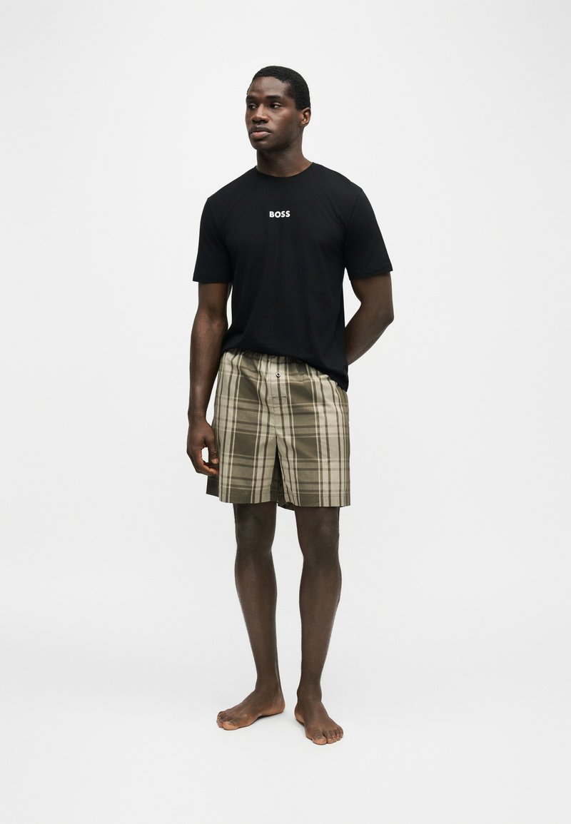 Homme debout pieds nus portant un T-shirt noir BOSS et un short beige à carreaux sur un fond blanc uni.