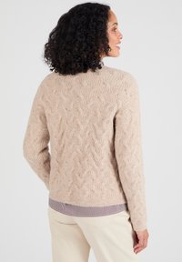 Donna con capelli ricci che sorride, indossando un maglione beige a treccia sopra una camicia a righe e pantaloni chiari, in posa di profilo su sfondo bianco.
