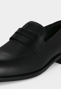 Mocassin en cuir noir avec tige texturée, détail de bride sur le dessus du pied et petit talon empilé sur fond blanc.