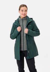 Groene waterdichte jas met capuchon, voorzien van een tweerichtingsrits en zijzakken, gedragen over een grijze fleece-top. Matte afwerking en een relaxte pasvorm.