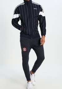 Námořnický pruhovaný sportovní kabát s bílými akcenty, vysokým límcem a zipovým uzávěrem, spárovaný se šedými tepláky s logem a žebrovanými manžetami.