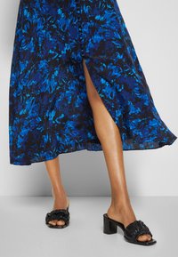 Femme portant une jupe midi fleurie bleue et noire avec une fente devant et des sandales noires à talons froncées sur un sol gris.