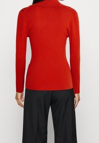 Czerwona prążkowana dzianinowa sweter z wysokim kołnierzem i długimi rękawami, o dopasowanym kroju i subtelnych akcentach na ramionach. Noszony z czarnymi spodniami.