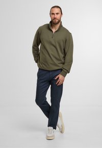 Felpa verde oliva con zip e maniche lunghe, abbinata a pantaloni blu scuro e sneakers bianche con dettagli testurizzati.