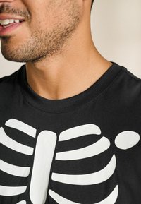 Sort t-shirt med et hvidt skelet ribben grafisk tryk foran. Rund halsudskæring, korte ærmer og blød bomuldsstruktur.
