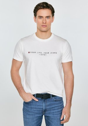 Młody mężczyzna w białej koszulce z napisem "Your Life. Your Jeans -1979-" i niebieskich dżinsach, stoi z jedną ręką w kieszeni.
