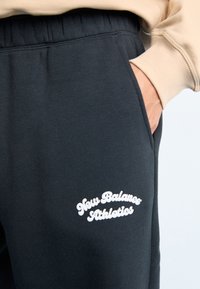 Pantalons de survêtement noirs en tissu doux, avec des poches latérales et un logo brodé blanc "New Balance Athletics" à l'avant.