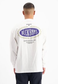 Wit shirt met lange mouwen, met een blauwe ovaalvormige afbeelding en de tekst "BLCKBNNS" op de achterkant, omringd door een gouden lauriertak ontwerp.