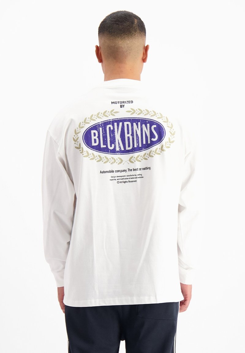 Wit shirt met lange mouwen, met een blauwe ovaalvormige afbeelding en de tekst "BLCKBNNS" op de achterkant, omringd door een gouden lauriertak ontwerp.