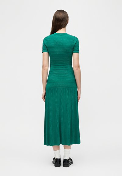 Femme de dos portant une robe courte à manches courtes texturée couleur sarcelle avec une jupe évasée, des chaussettes blanches et des chaussures noires sur un fond blanc uni.