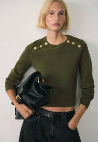 Maglione verde a costine con bottoni dorati sulle spalle, abbinato a una borsa in pelle nera, indossato con jeans scuri a vita alta.
