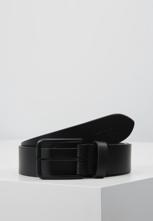 Ceinture en cuir noir avec boucle rectangulaire noire mate enroulée sur un piédestal blanc sur un fond gris clair uni.