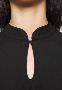 Svart topp med rundad krage och en nyckelhålsöppning i mitten. Slät tygtextur utan synliga mönster eller utsmyckningar.