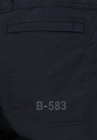 Svart tyg med ett rutmönster som har en bakficka. Synlig är en broderad detalj med texten "B-583."