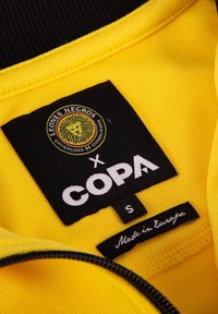 Gele kleding binnenkant met een zwarte rits. Het label bevat een leeuwlogo, de tekst "LEONES NEGROS," "COPA," maat "S," en "Gemaakt in Europa."